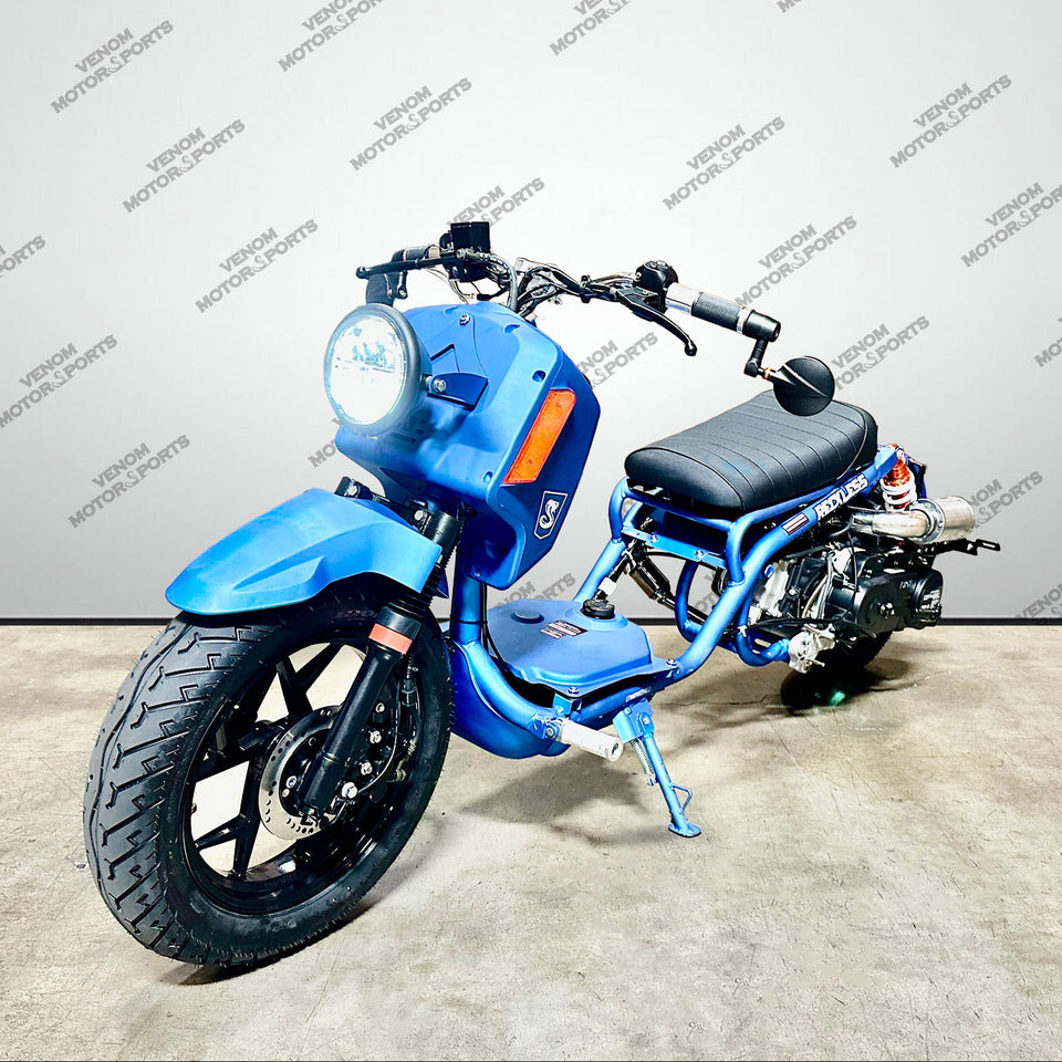 A blue gas ruckus style motorcycle with black wheels, a black seat, and orange signal lights, photographed indoors against a light gray wall with repeated watermark text.